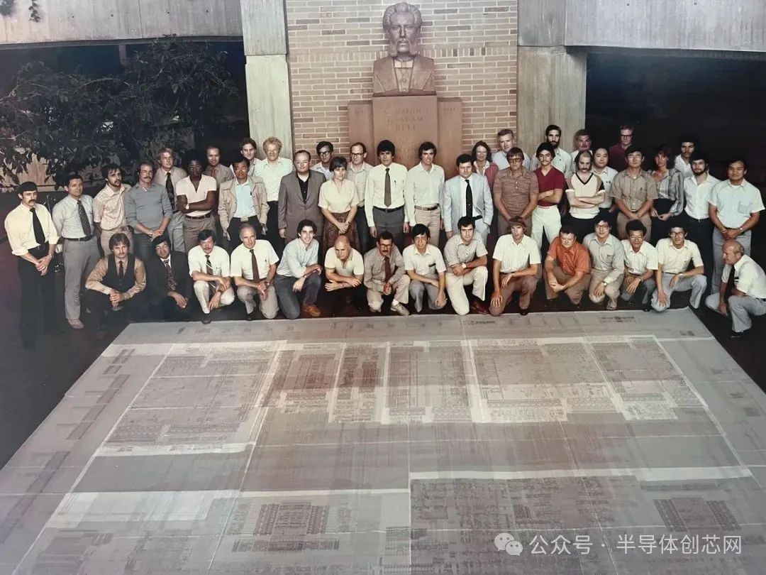 Group posing around a large electronic circuit schematic, with a sculpture in the background.